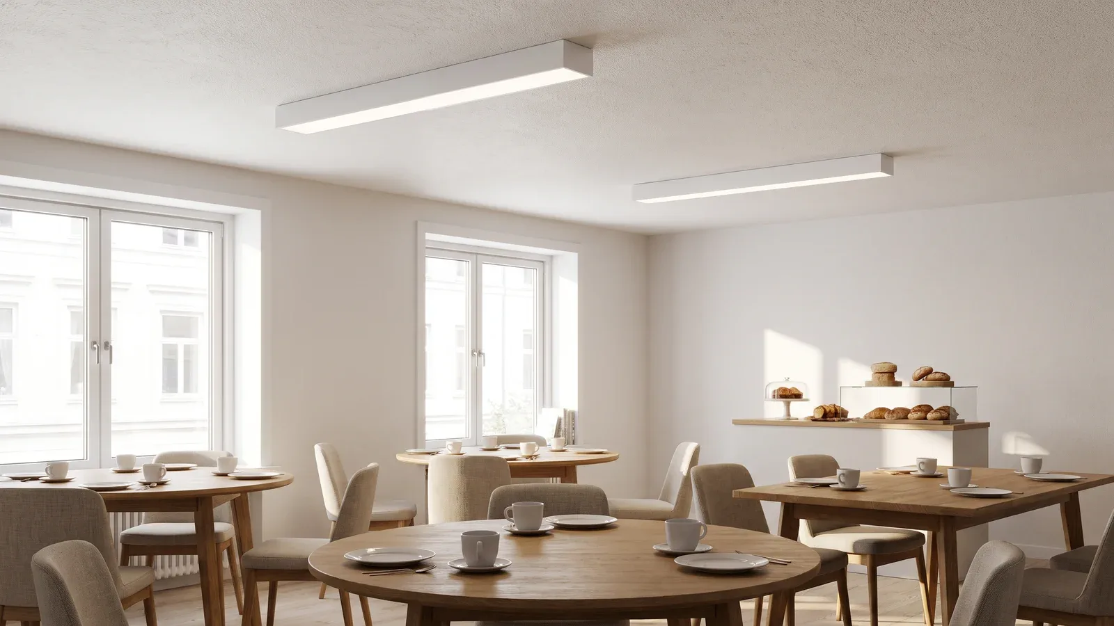 Salón de desayunos de hotel con luminarias lineales blancas de superficie sobre las mesas y luz natural.